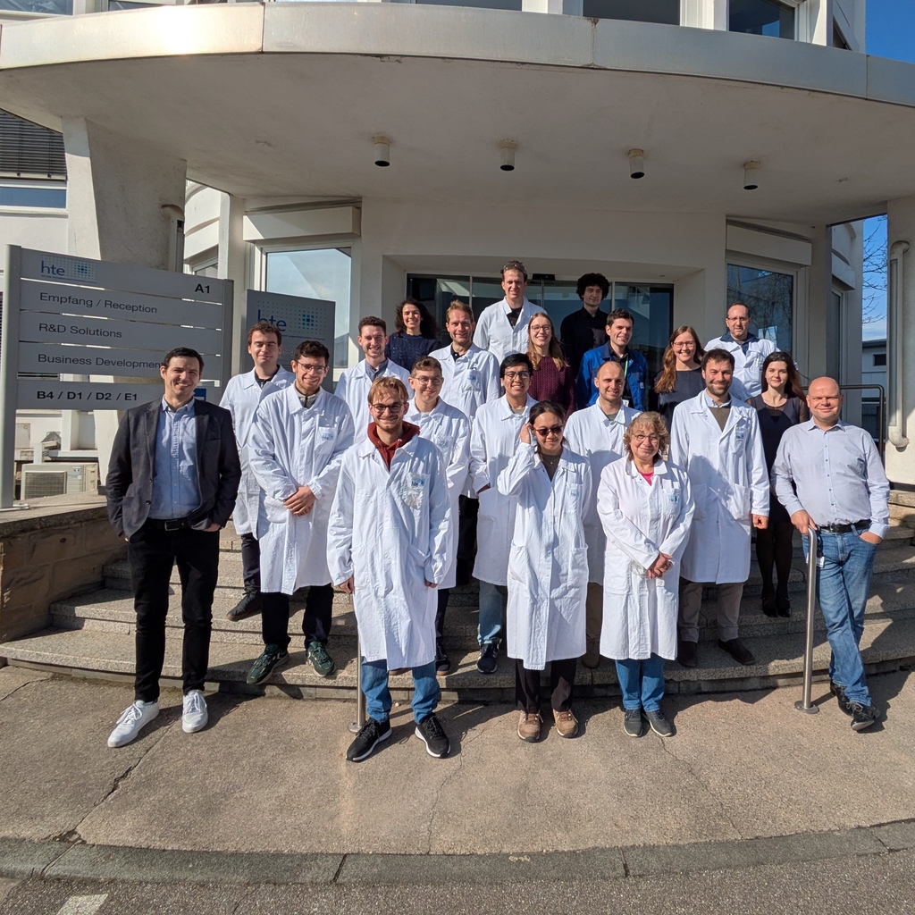 Gruppe von Studierenden und Mitarbeitenden vor einem Gebäude, einige tragen weiße Laborkittel.